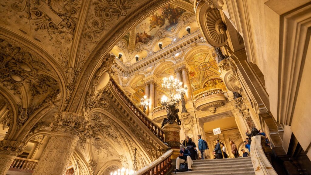 "Grand staircase inside the Palais Garnier in Paris, showcasing opulent chandeliers, ornate columns, and a richly decorated ceiling with classical frescoes, as visitors explore the historic opera house."